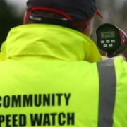 Person in a high-vis jacket reading "Community Speed Watch" using a radar speed gun outdoors.