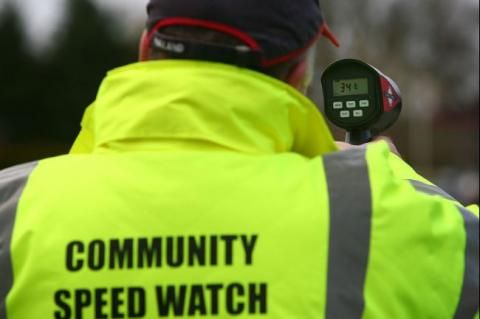 Person in a high-vis jacket reading "Community Speed Watch" using a radar speed gun outdoors.