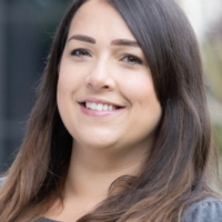 Smiling woman with long brown hair wearing a grey top, standing outdoors.