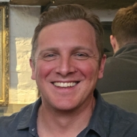 A smiling man in a blue shirt sits at a wooden table in a cosy café with shelves and bread in the background.
